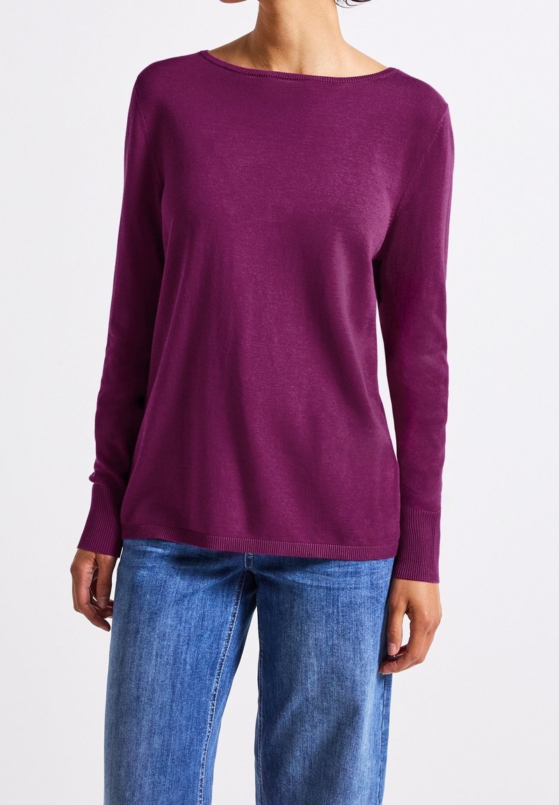 Personne portant un pull violet à manches longues associé à un jean en denim bleu sur un fond blanc uni.