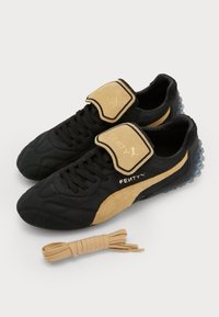 Black and tan sneakers with textured leather, a padded collar, velcro strap, and gold branding accents. Includes a bundled beige shoelace.