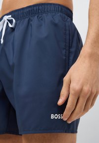 Tamsiai mėlyni plaukimo šortai su lygiu paviršiumi, elastine juosmens juostele ir balta raišteliu, ant apatinių dešiniosios kojos dalių esantis baltas „BOSS“ logotipas.