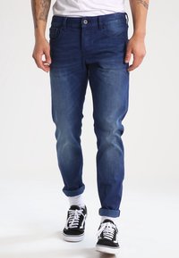 Vaqueros de mezclilla azul oscuro con un corte slim, un desvanecimiento sutil y puños enrollados. Combinados con zapatillas de deporte en blanco y negro, y una camiseta blanca lisa.