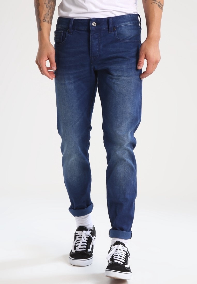 Vaqueros de mezclilla azul oscuro con un corte slim, un desvanecimiento sutil y puños enrollados. Combinados con zapatillas de deporte en blanco y negro, y una camiseta blanca lisa.