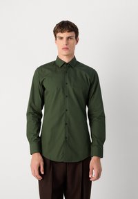 Cămașă butonată de culoare verde închis, cu guler clasic, mâneci lungi, nasturi negri și o textură netedă. Purtată cu pantaloni maro.