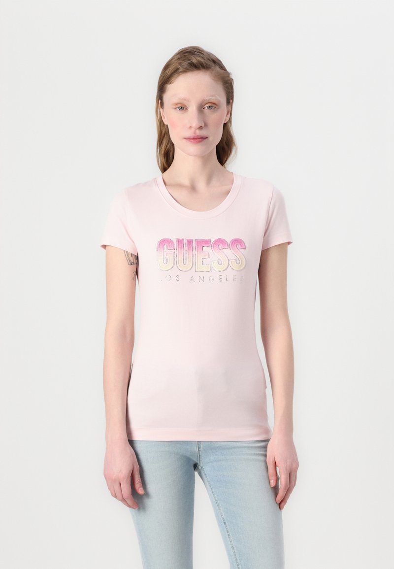 T-shirt rose clair à manches courtes en matériau doux, arborant un logo "GUESS" en dégradé avec des accents scintillants et un col rond.