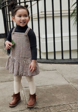Robe tablier marron à carreaux avec ourlet froncé, associée à un haut à manches longues noir, des collants en tricot crème et des bottines marron.