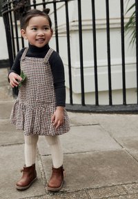 Vestito salopette marrone a quadri con orlo arricciato, abbinato a una maglia nera a maniche lunghe, calze in maglia color crema e stivaletti marroni.