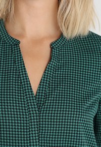 Grön houndstooth-mönstrad topp med V-ringning, som har ett texturerat tyg och subtila rynkor vid ringningen.