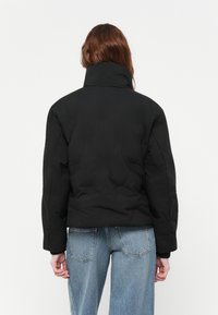 Svart kort pufferjakke med høy krage, teksturert stoff og ribbede mansjetter. Bært med lyseblå vide jeans.