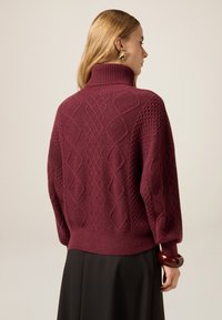 Vinröd stickad tröja med hög krage, med texturerade diamantmönster och ribbade ärmslut. Bärs med en mörk kjol och accessoarer.