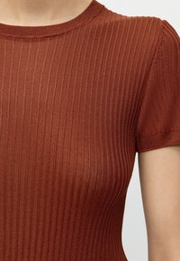 Gros plan d’une personne portant un haut moulant à manches courtes en maille côtelée couleur rouille sur un fond neutre.