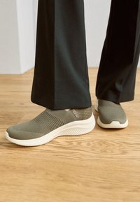 Sneaker slip-on in tessuto a maglia verde oliva con dettagli testurizzati, suole bianche e design a profilo basso, abbinate a pantaloni neri.
