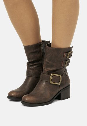 Bottines en cuir marron avec fermeture éclair sur le côté, deux boucles décoratives près du haut, et un talon bas épais, portées sur des jambes nues.