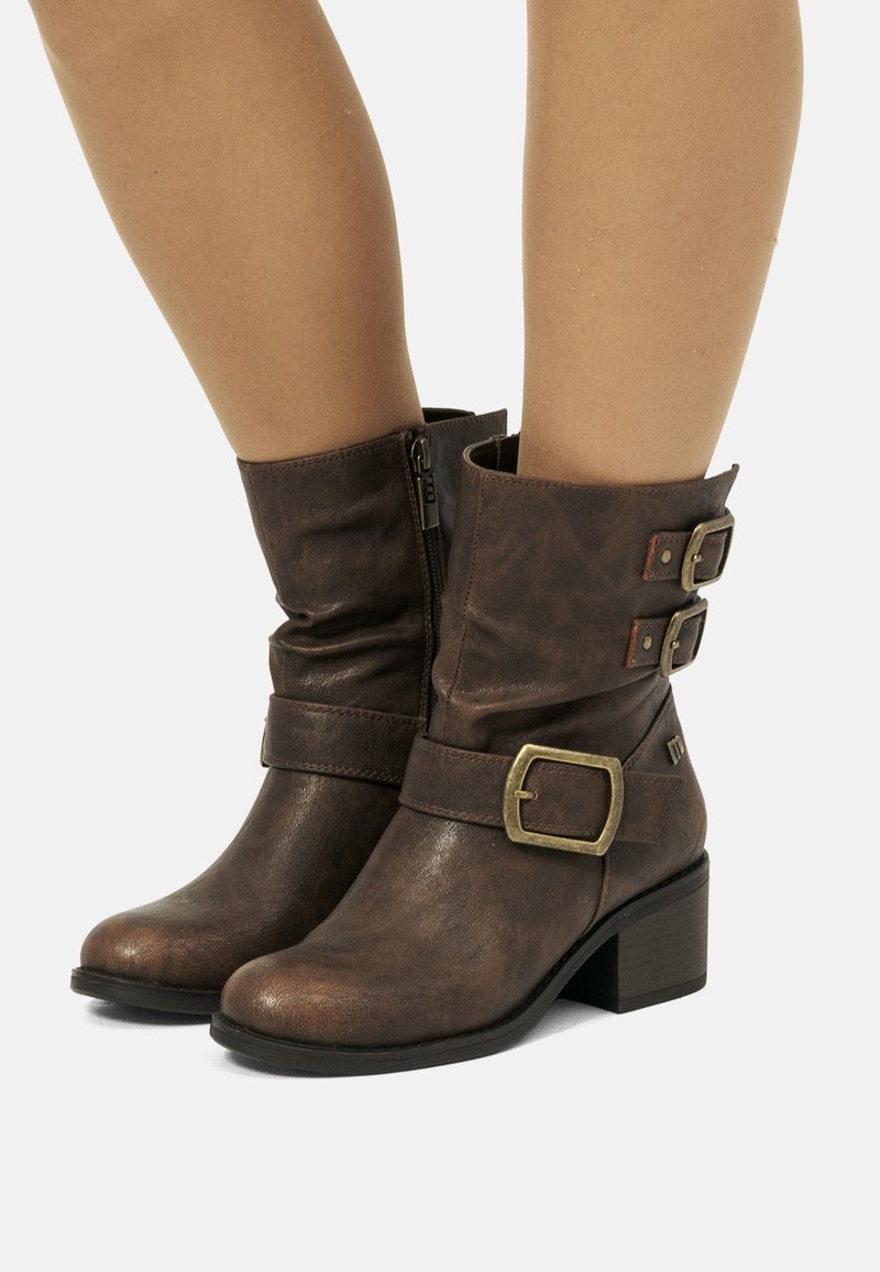 Stivali ankle in pelle marrone con cerniera laterale, due fibbie decorative vicino alla parte superiore e un tacco basso e tozzo, indossati su gambe nude.