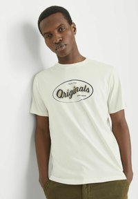 Crème kleurige katoenen t-shirt met korte mouwen, met een marineblauwe ovale "Originals" grafische print over de borst. Eenvoudig, casual ontwerp.