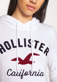 Jeune personne portant un sweat à capuche blanc avec un texte noir "Hollister California" et un logo d'oiseau rouge à l'avant.