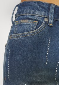 Tamno plave traperice sa prilagođenim dizajnom, svijetlim oštećenjima i srebrnim zakovicama duž bočnog šava. Vidljiv Pepe Jeans logo.