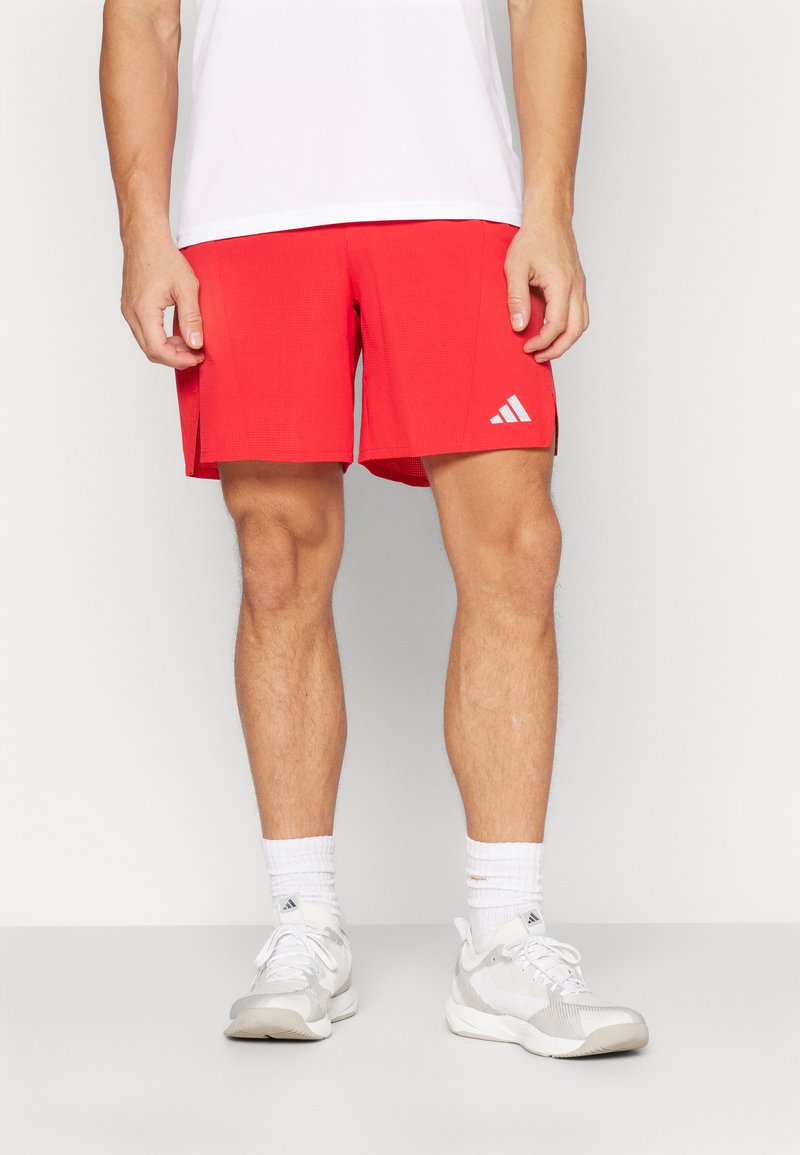 Shorts de sport rouges avec une surface texturée, des détails de logo blancs sur le côté et des fentes sur les côtés pour faciliter les mouvements. Associés à des baskets grises.