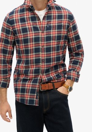 Mężczyzna ubrany w granatową, czerwoną i białą koszulę w kratę na białym t-shircie, ciemne dżinsy, brązowy skórzany pasek i złoty zegarek na rękę.