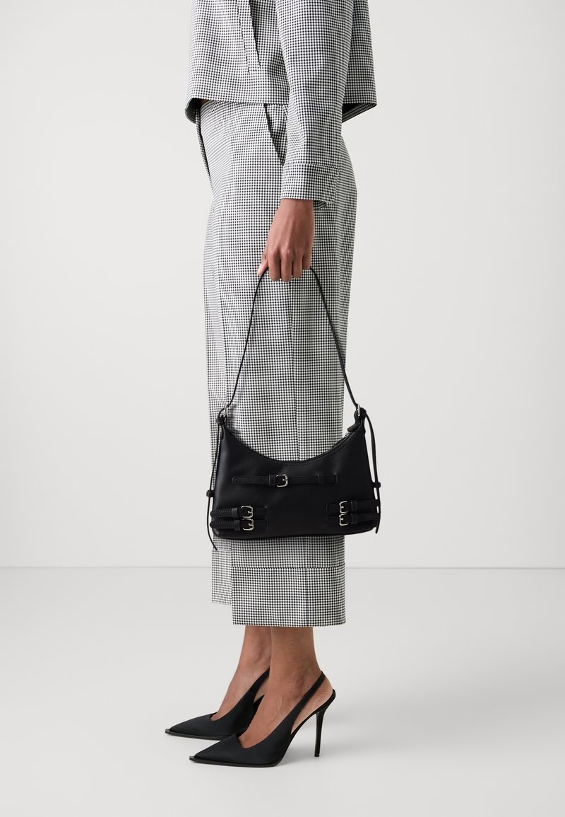 Personne en tailleur-pantalon à carreaux tenant un petit sac à main noir avec des boucles, portant des escarpins slingback noirs à talons hauts et pointus.