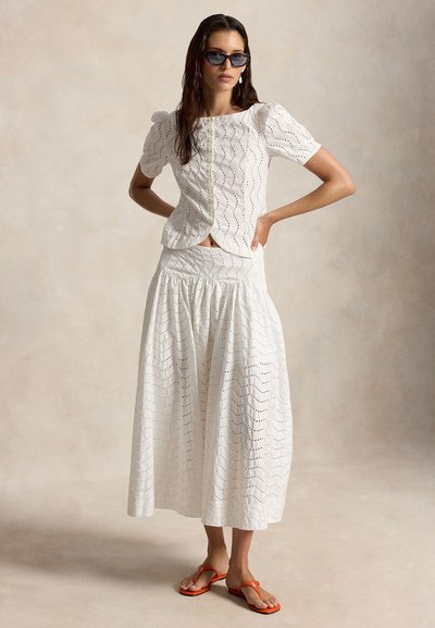Femme portant un chemisier blanc en broderie anglaise avec des manches bouffantes, une jupe longue assortie, des lunettes de soleil noires et des sandales orange, debout les mains sur les hanches.