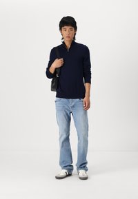 Suéter de cremallera azul marino, pantalones vaqueros claros y zapatillas grises. Una pequeña bolsa negra está colgada del hombro. Tejido de punto suave, corte informal.