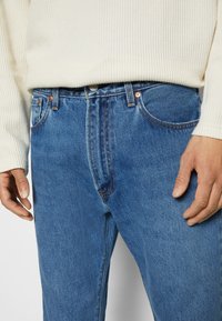 Gros plan sur une personne portant un jean en denim bleu et un pull crème côtelé, les mains détendues le long du corps.