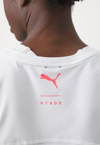 Biała koszulka sportowa z różowym logo Puma i napisem "HYROX" z tyłu, o gładkiej tkaninie i okrągłym dekolcie.