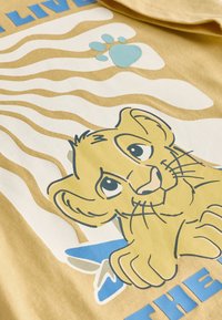 Sárga pamut póló stilizált oroszlán grafikával, kék mancslábbal és hullámos csíkokkal bézs és krémszínű háttéren.