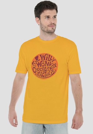 Mann trägt ein gelbes T-Shirt mit einem roten, runden „Willy Wonka The Chocolate Factory“-Design und schaut zur Seite.