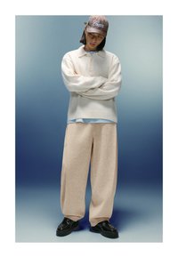 Osoba v bežových širokých nohaviciach, krémovom svetri, svetlomodrej košeli, hnedom károvanom klobúku a čiernych topánkach stojí so založenými rukami na gradientnom modrom pozadí.