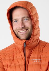 Oranje pufferjack met capuchon, met een quilted ontwerp en een voorrits. Branding met "SWEDEMOUNT" in het wit. Glad textuur.