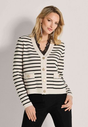 Vrouw draagt een zwart-wit horizontaal gestreepte cardigan met gouden knopen en een zwarte broek, staand tegen een effen lichte achtergrond.