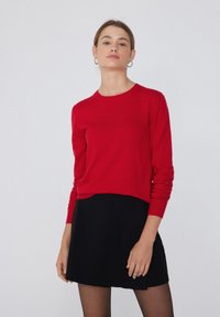 Pull rouge à manches longues en matière douce avec un col rond, associé à une jupe noire. Texture lisse avec une coupe décontractée.