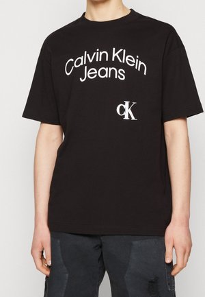 Osoba nosząca czarny t-shirt Calvin Klein Jeans z białym napisem logo i ciemne spodnie z przetartą łatą na lewej nodze.