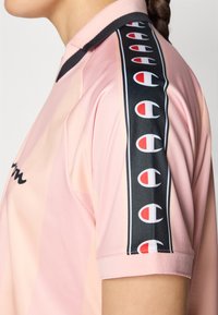 Heled roosa spordijerseys, millel on must küljeriba, kaunistatud korduvate logodega punases ja valges. Lühikesed varrukad, nähtav krae disain.