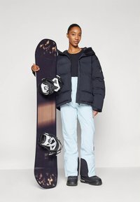 Žena v černé bundě, světlomodrých sněhových kalhotách a černých botách drží snowboard s vázáním proti jednolitému pozadí.