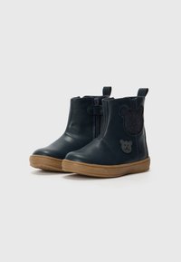 Botas de tobillo de piel azul marino con cremallera lateral, suelas de goma color marrón y un acento de oso brilloso en el lado exterior.