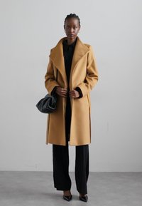 Camel-colored coat with a structured collar, long sleeves, and open front, paired with black trousers and a black clutch handbag.