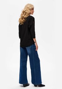 Blonde woman with wavy hair wearing a black long-sleeve shirt, wide-leg blue jeans, and black heeled shoes, standing facing sideways.