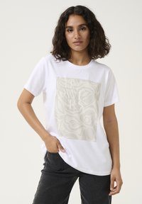 T-shirt blanc en coton présentant un grand motif floral subtil en beige et le mot "FLEURS" brodé en blanc sur le devant.