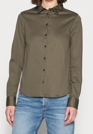 Femme portant une chemise olive à manches longues boutonnée et un jean en denim bleu clair, debout devant un fond uni.