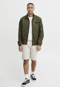 Olive green lightweight jacket with a zip closure, elastic cuffs, and a small pocket. Worn over a white t-shirt and paired with beige shorts and black sneakers.