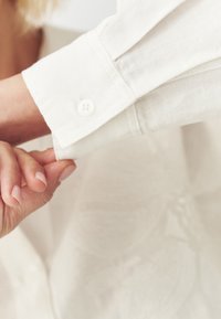 Světlé béžové manžety košile s hladkým povrchem, se single bílým knoflíkem. Ruka uchopující manžetu, ukazující jasné detaily látky.