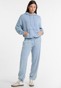 Gaiši zils kapučis ar regulējamu auklu, priekšējo kabatu un brīvu piegriezumu, apvienots ar saskaņotām šaurajām sporta biksēm. Mīksta tekstūra un ikdienišķs dizains.