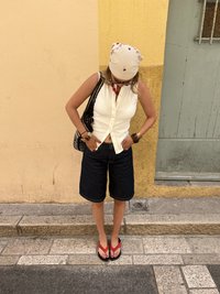 Femme portant une chemise blanche sans manches boutonnée, un short noir, des tongs rouges et un bandana blanc, regardant vers le bas sur un trottoir.