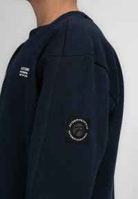 Felpa blu navy con collo rotondo e finitura testurizzata. Presenta un'etichetta con logo sulla manica e testo ricamato sul petto.