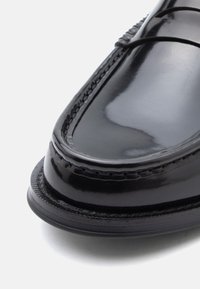 Mocassins en cuir noir à finition brillante, avec des coutures décoratives le long des bords et un design à bout arrondi.