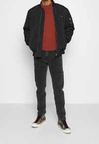 Chaqueta bomber negra con textura suave, camiseta de cuello redondo roja, pantalones cargo oscuros y zapatillas de deporte con patrón multicolor.