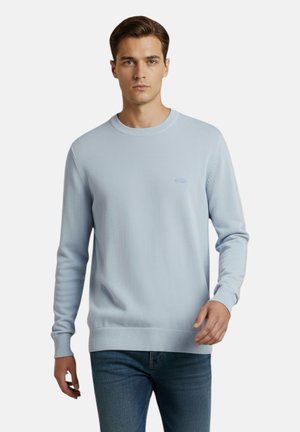 Uomo che indossa un maglione a maniche lunghe in maglia azzurro chiaro con scollo a girocollo e jeans blu su uno sfondo bianco semplice.