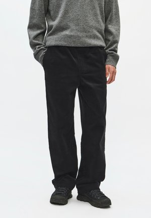 Personne portant un pantalon en velours côtelé noir, des chaussures de randonnée noires et un pull gris à manches longues, les mains dans les poches, debout sur un fond blanc uni.
