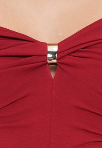 Top rojo con un diseño retorcido en el escote, que presenta un acento en forma de anillo en tono dorado. Tejido suave, forma ajustada y corte en forma de V en el escote.